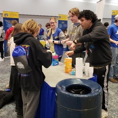 Three chemistry club students talking to two students and performing an experiment while standing at table during a middle school outreach fair.