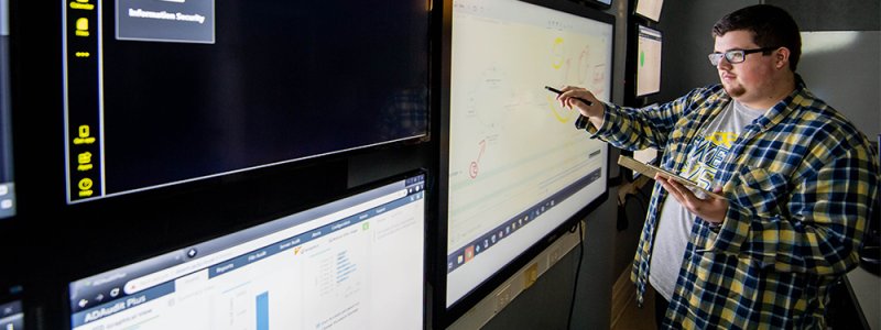 man stands in front of a wall of computer screens analyzing data