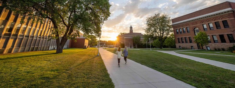 Shot of two students walking on campus with blue skies and the sun coming up over the buildings.