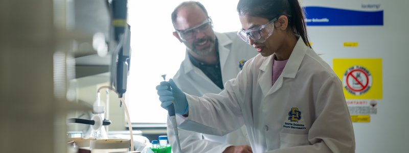 A professor watching a student perform a scientific experiment with beakers.