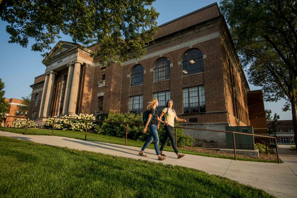 Two students walking on sidewalk in front of a residence hall.