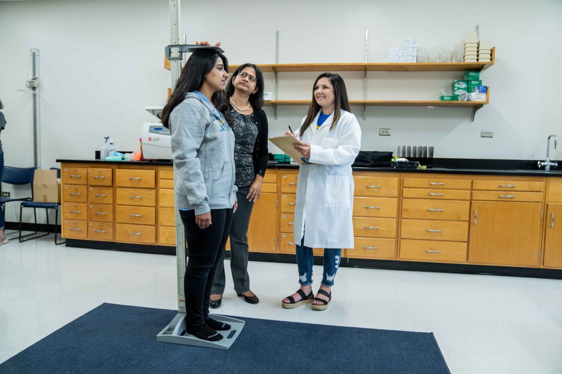 Student being measured by height by a student in a white lap coat and a preceptor.