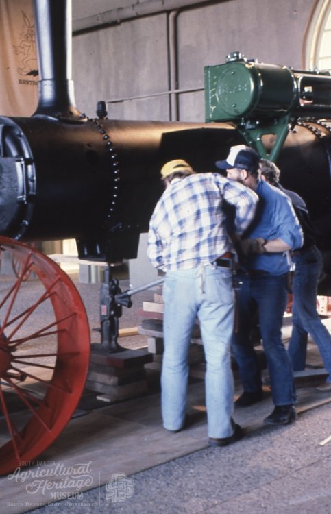 A group of men are reattaching the front wheels on the steam engine.  The tractor is black and the wheels are red.  There are three men working on the tractor.