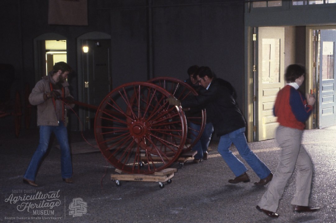 Two men moving a section of a steam engine tractor into the building.  This section has two red wheels attached.  There is also a women walking through the side of the photo.