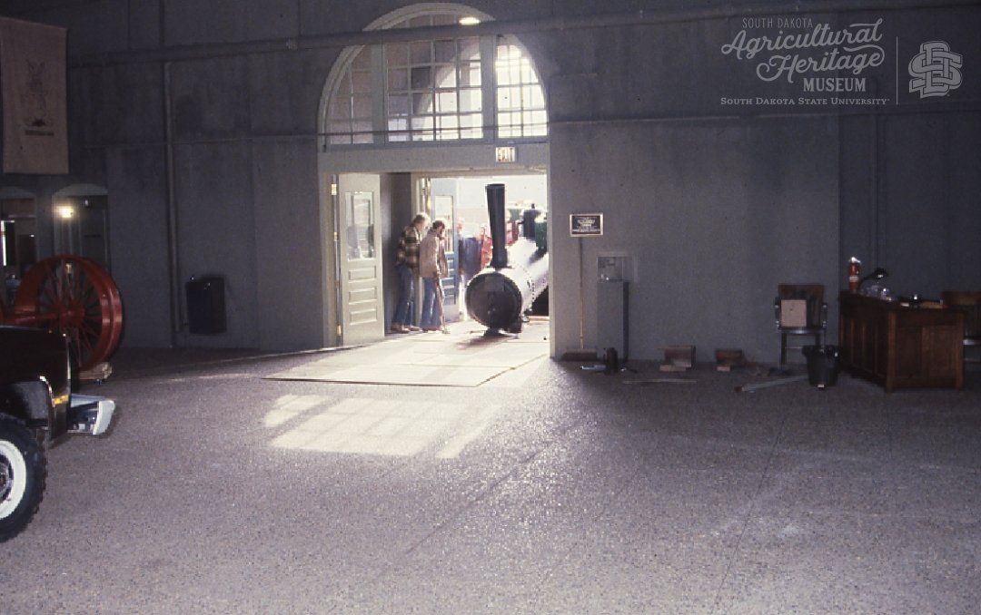 A group of men moving the steam block of the steam engine tractor into the museum.  The walls and floors are gray and the doorway they are moving it through has a section of curved windows at the top.