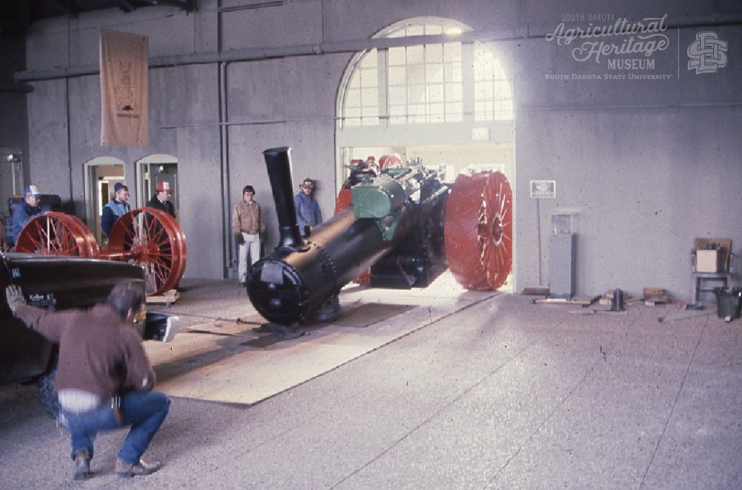 Moving the steam engine into the museum.  There is plywood laid beneath the engine to prevent the floor from damage.  The front wheels are taken off the engine so it can fit through the doorway.  The tractor is black with red wheels.  A group of men are helping move the tractor through the door.