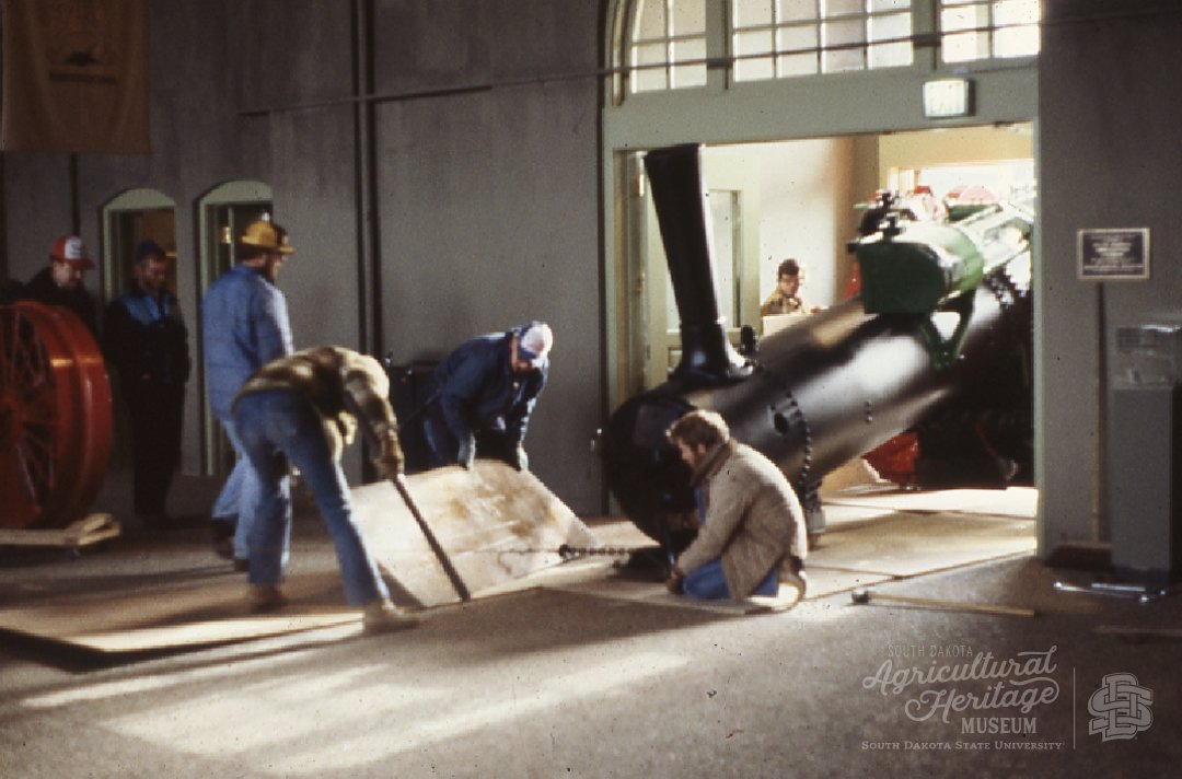 Moving the steam engine into the museum.  The walls and floors are gray and the steam engine tractor is black.  there are four men around the front of the tractor that are using items to slide the engine in.